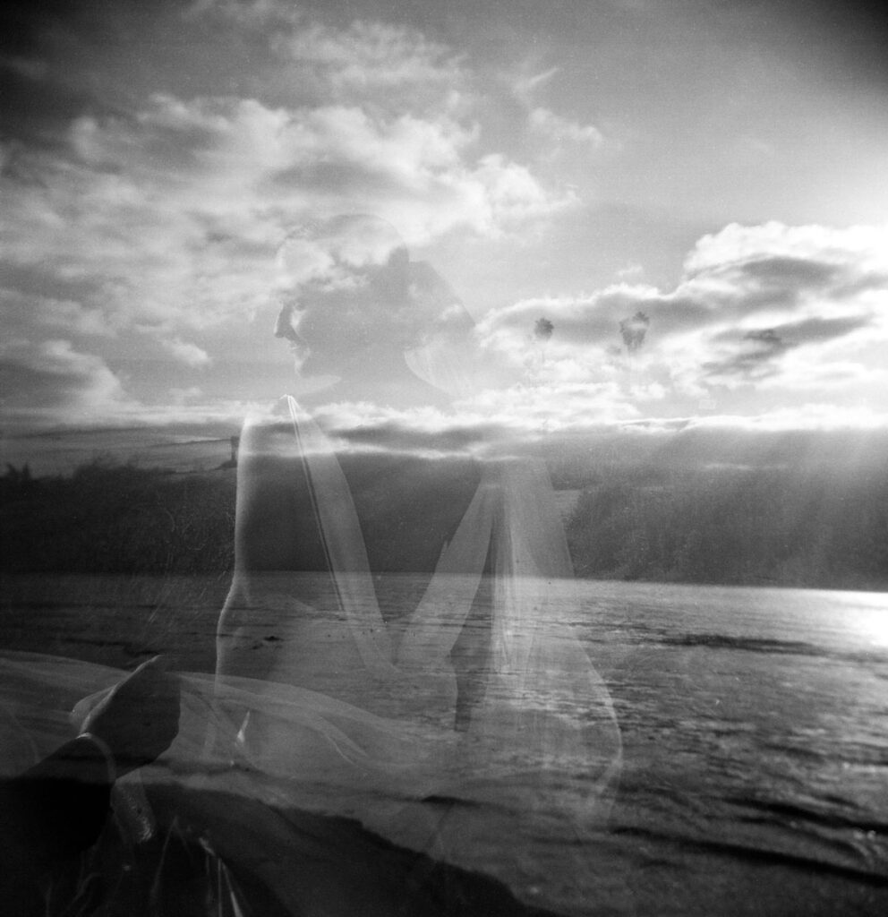 Black and white Holga photo of a ghostly double-exposure of the bride's veil over the ocean