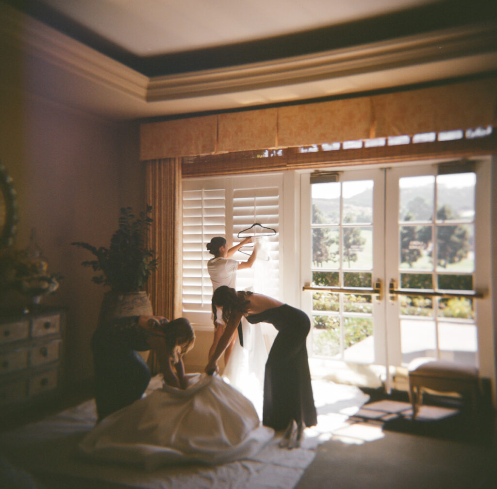 Bridesmaids helping bride into her gown by the window at Del Mar Country Club.
