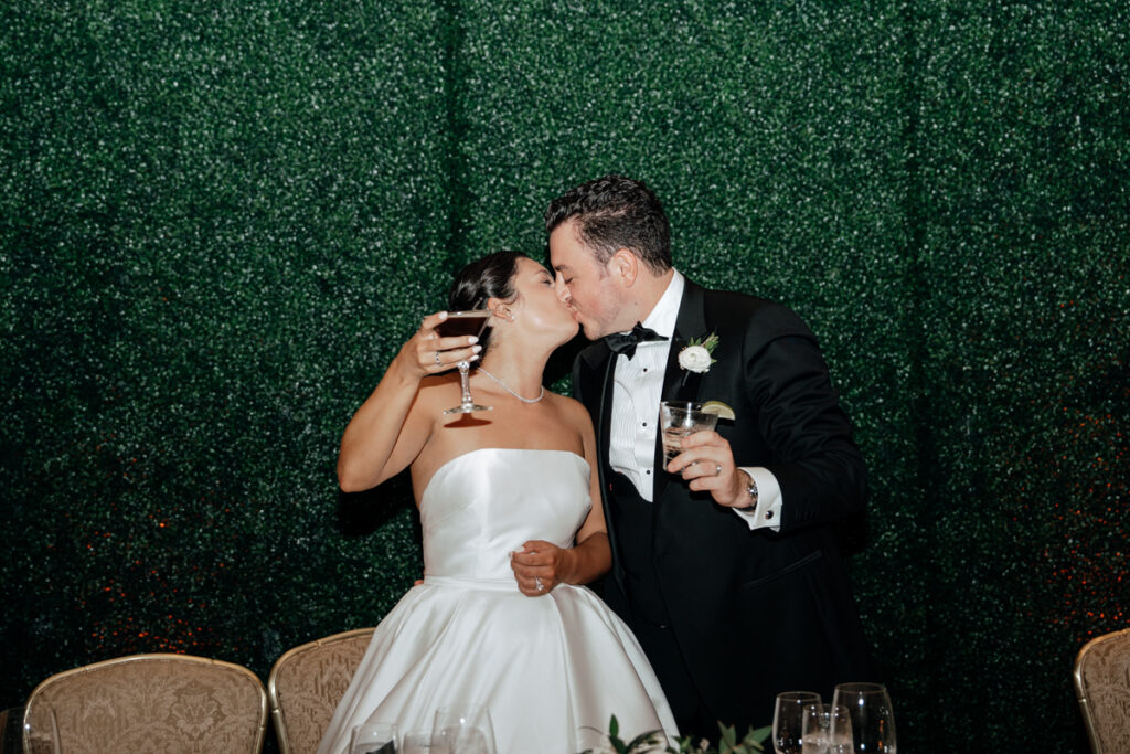 Bride and groom sharing a toast and kiss in front of greenery wall at Del Mar Country Club.