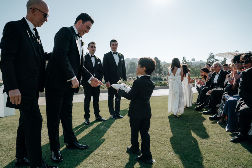 Young ring bearer in suit handing rings to groom during Del Mar Country Club wedding ceremony.