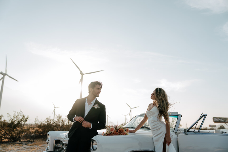 Bride and groom sharing a candid moment beside classic convertible in the desert