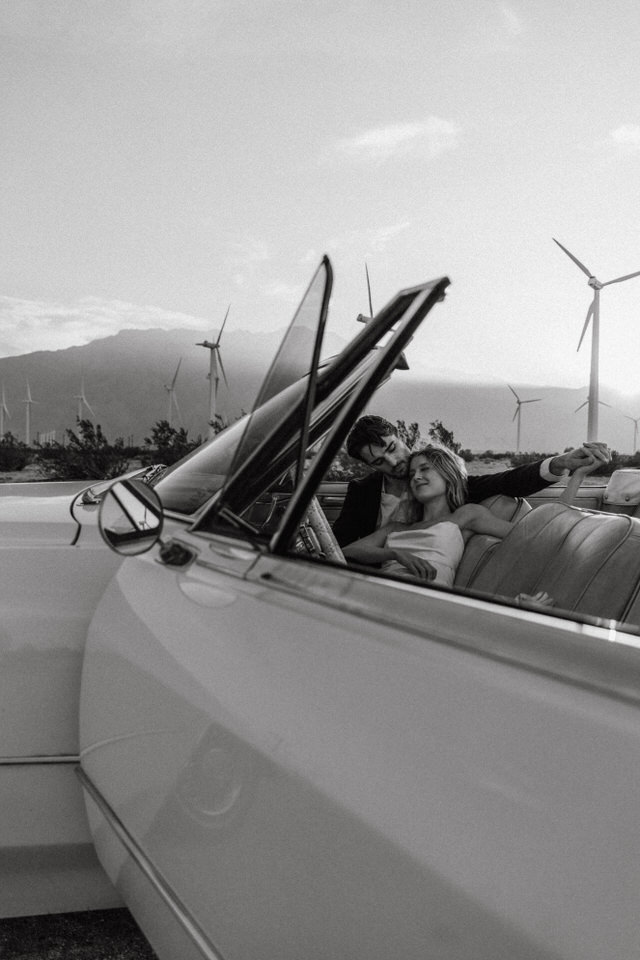 Black and white wedding portrait of couple relaxing in vintage convertible in desert landscape