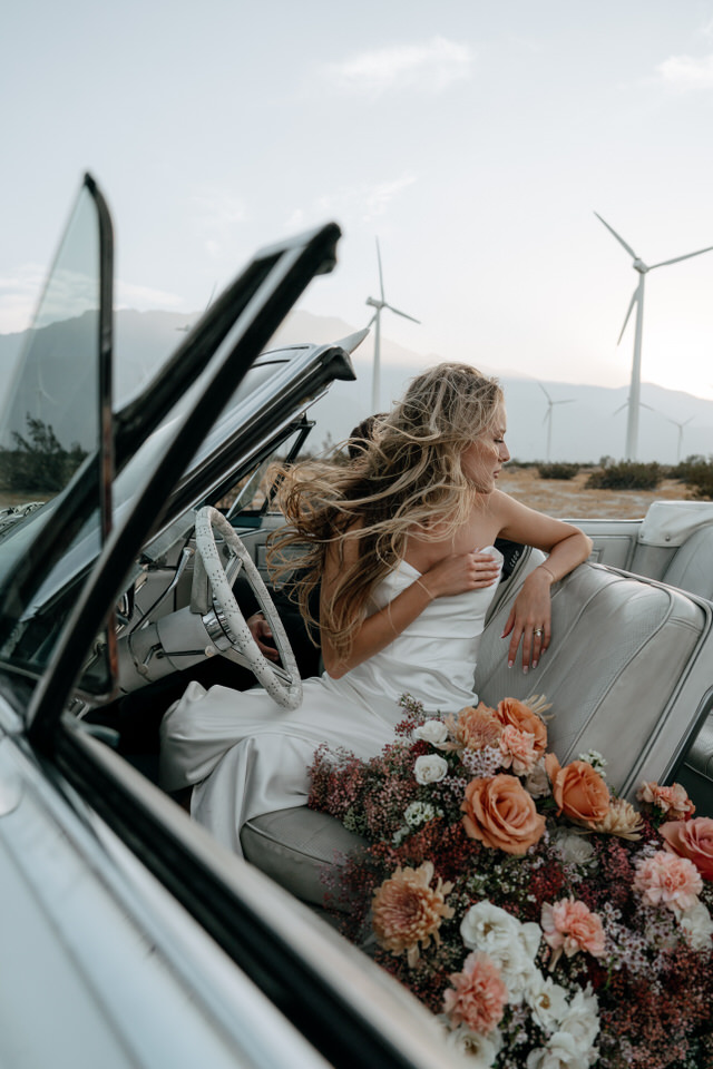 Bride leaning back in vintage convertible with colorful bouquet at Palm Springs wind farm