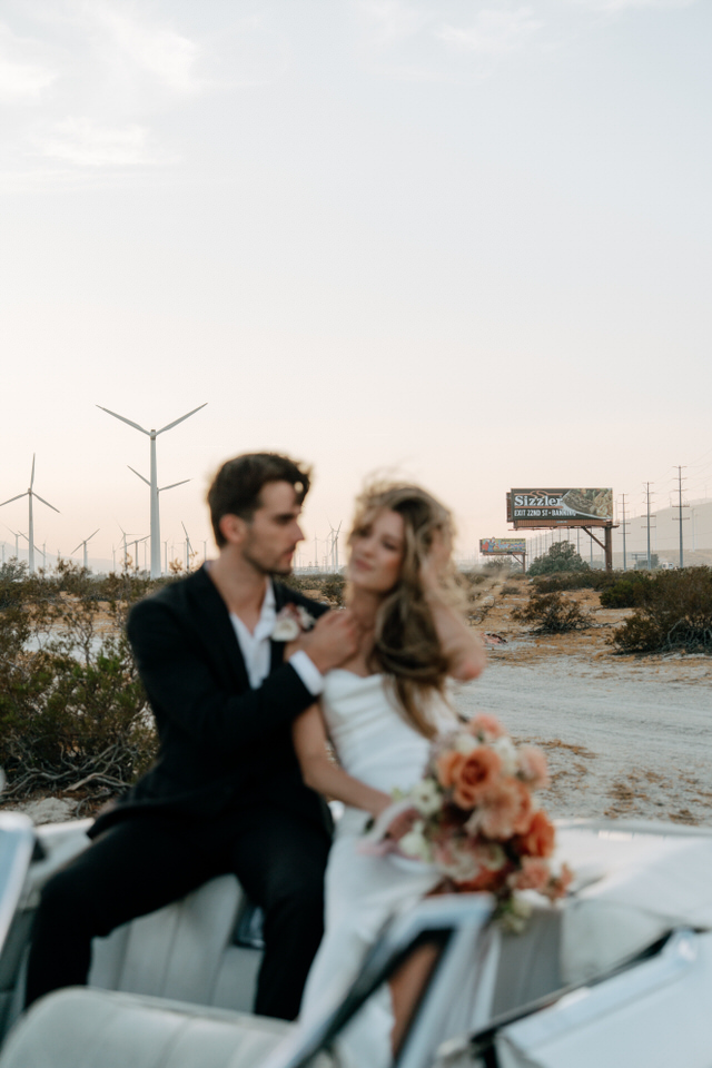Romantic wedding portrait of couple sitting on convertible car with bouquet at Palm Springs windmills