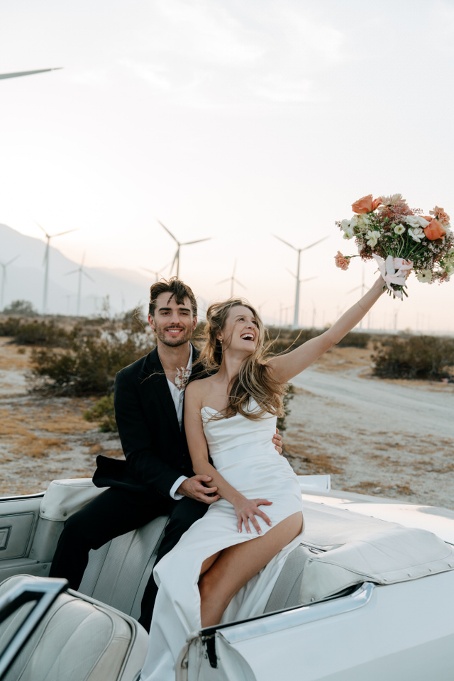 Couple celebrating their elopement in convertible, bride holding bouquet high in Palm Springs desert