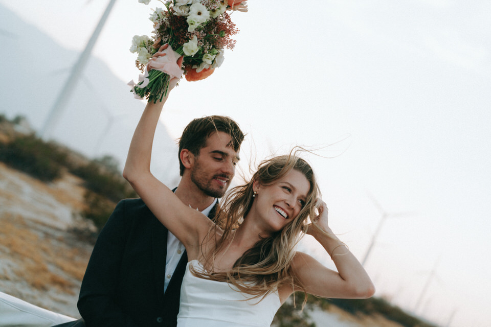 Bride smiling and lifting bouquet while groom embraces her during candid Palm Springs windmill photos