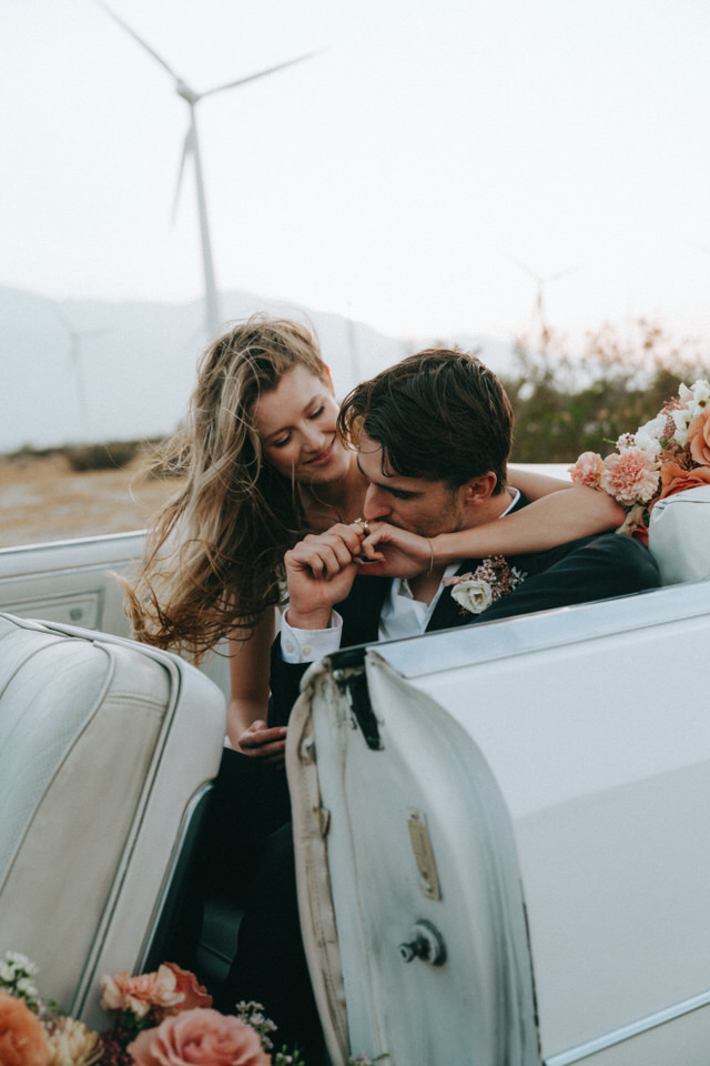 Bride leaning over car door to kiss groom during candid wedding photos in desert setting