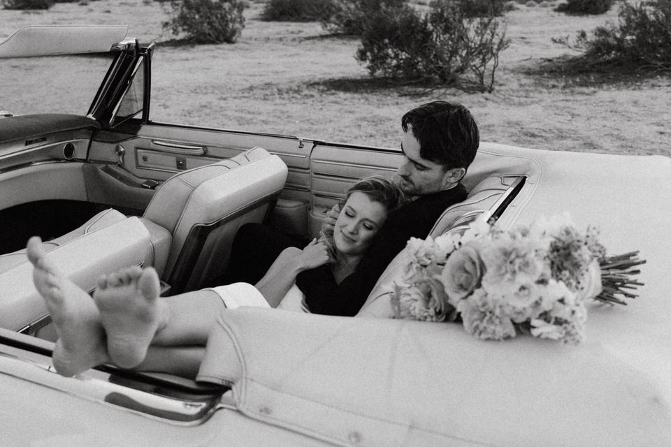 Black and white candid wedding photo of couple barefoot and relaxed in vintage convertible with bouquet nearby