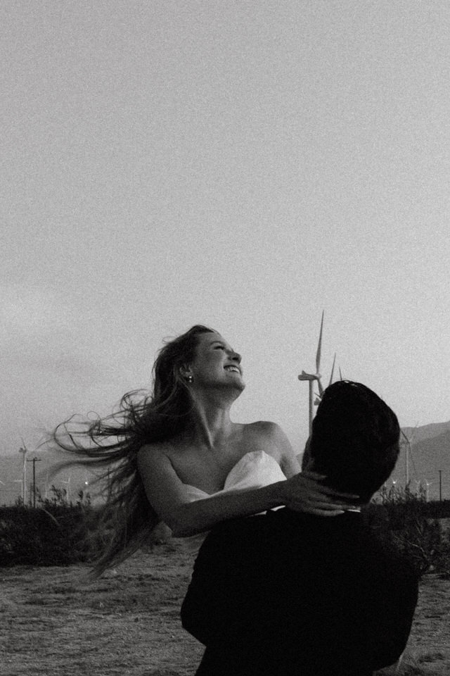 Black and white candid photo of groom lifting bride with turbines in the distance