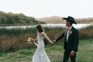 Bride and groom walking hand in hand along the water's edge at Alisal Ranch, groom in a black cowboy hat, bride holding her bouquet