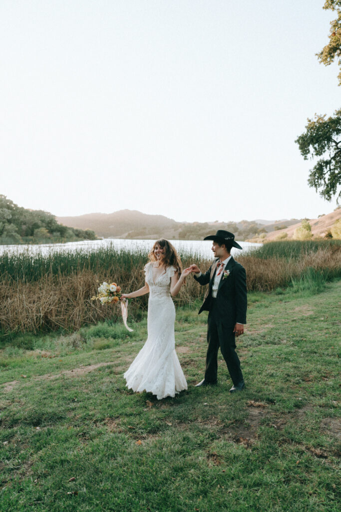 Groom in a cowboy hat and bride laughing together in an open field by the lake