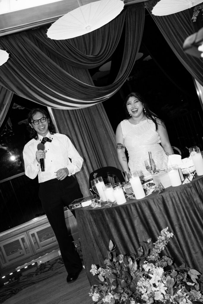 Black and white photo of the groom giving a toast at the sweetheart table, paper parasols overhead