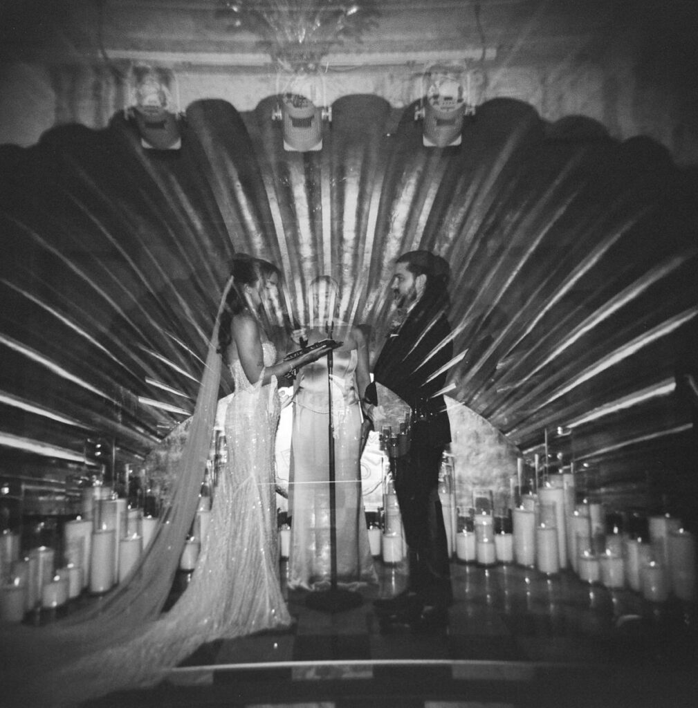 Double exposure of the couple exchanging vows in front of a scalloped, sunburst-lit stage backdrop lined with candles