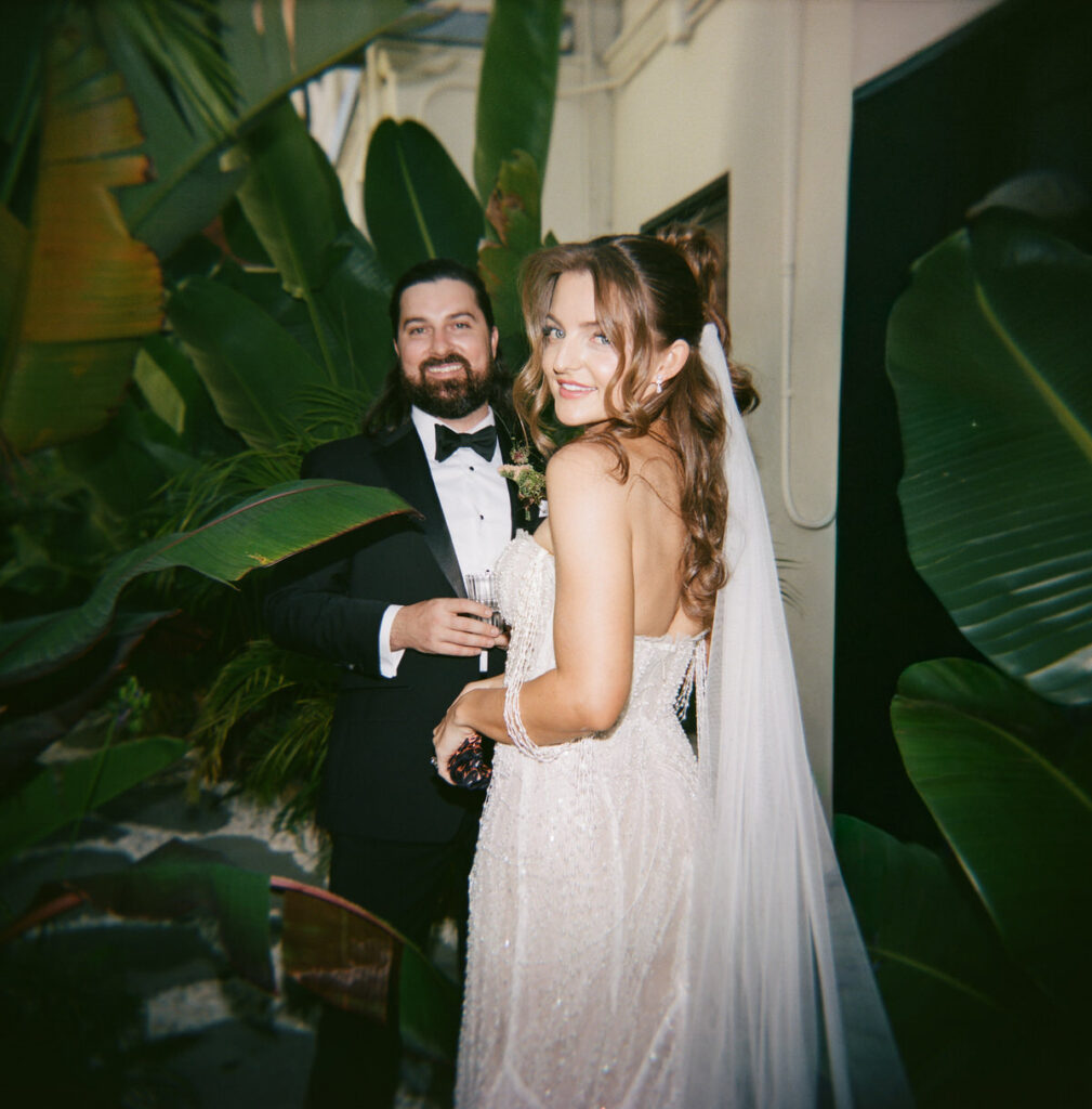 The bride looking back over her shoulder among banana leaf plants, veil flowing, groom visible behind her